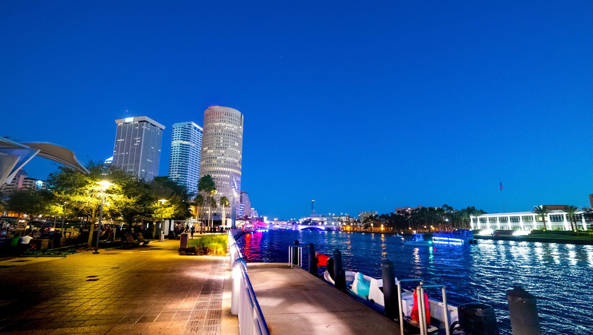 Illuminated city skyscrapers at night reflecting in a river from a lit waterfront walkway.