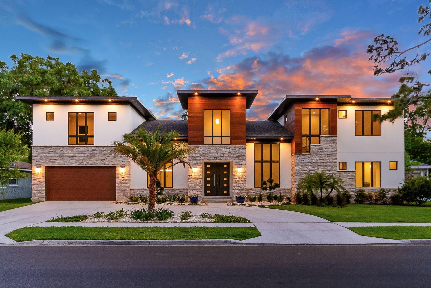 Modern two-story luxury home with stone and wood accents under a colorful sunset sky.